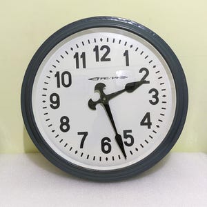 Könnte beinhalten: Eine graue Wanduhr aus Metall mit schwarzen Zahlen und Zeigern. Das Zifferblatt der Uhr ist weiß und trägt den Schriftzug "CRE/A".