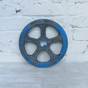 Op de afbeelding: Een cirkelvormig metalen handwiel met een ontwerp met zes spaken en een centrale zeshoekige opening. Het wiel heeft een verweerd uiterlijk en vertoont een combinatie van origineel grijs metaal en afgebladderde, versleten blauwe verf.