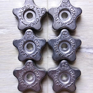 Peut inclure: Six boutons argentés en forme d'étoile, disposés en deux rangées de trois. Chaque bouton comporte un trou central et du texte en cercle, dont "OTKP" et "PB6-2". Ils sont probablement destinés à un usage industriel ou mécanique.