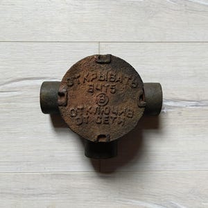 Puede incluir: Una caja de conexiones de hierro fundido oxidada con cuatro aberturas. La caja tiene el texto "SK KPDISAB 3LTS" y "OK KHOLINE 011 GETYN" grabado en la parte superior.