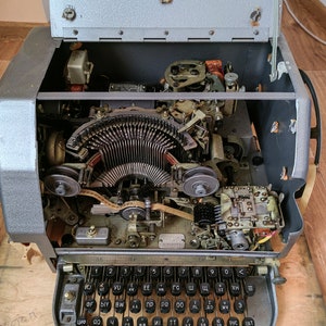 May include: A vintage typewriter with the top cover open, revealing the inner workings of the machine. The typewriter has a gray metal body and a black keyboard with Cyrillic lettering. The keyboard has keys labeled with numbers, letters, and symbols.