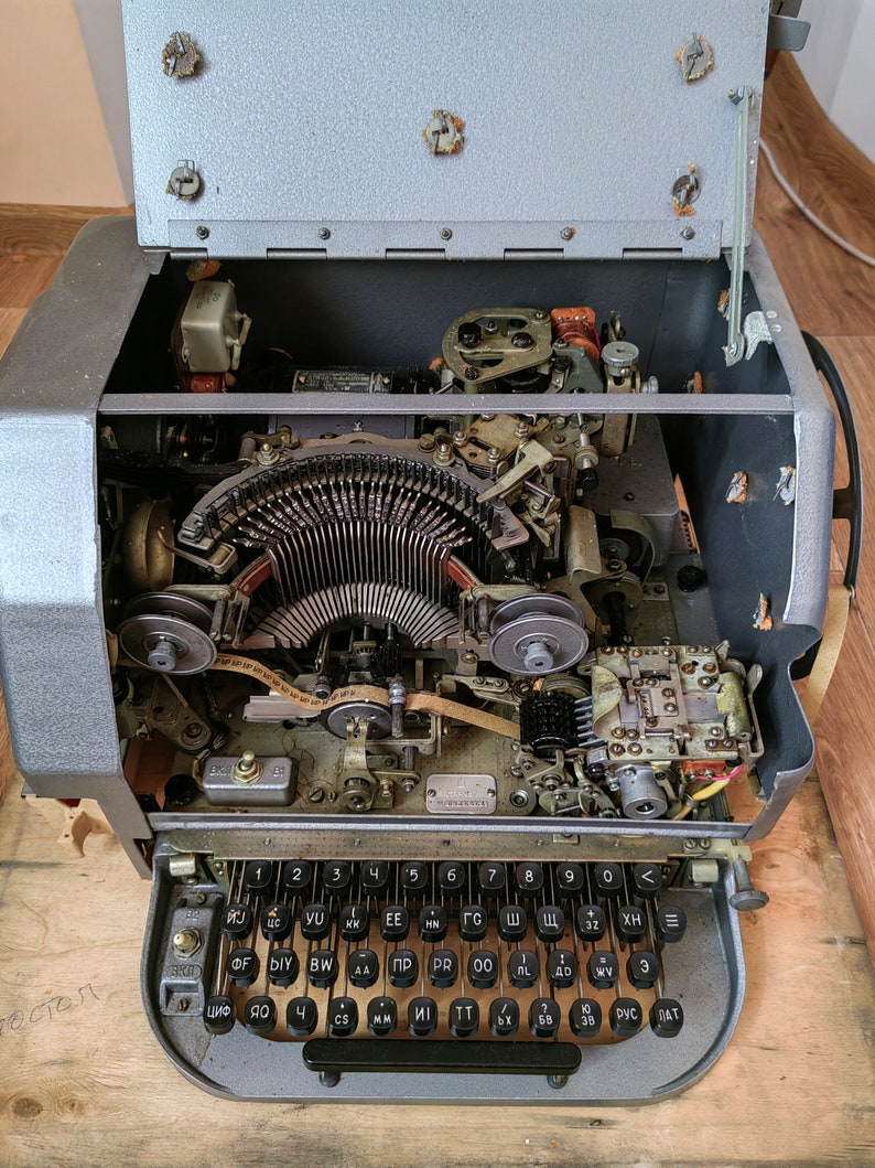 May include: A vintage typewriter with the top cover open, revealing the inner workings of the machine. The typewriter has a gray metal body and a black keyboard with Cyrillic lettering. The keyboard has keys labeled with numbers, letters, and symbols.