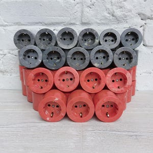 May include: A collection of vintage electrical outlets. The outlets are arranged in rows, with a mix of red and gray colors. Each outlet has three holes for plugs and a circular design.