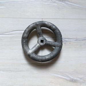 Könnte beinhalten: Ein großes, rundes, graues Metallrad mit drei Speichen und einem quadratischen Loch in der Mitte. Das Rad hat eine raue, strukturierte Oberfläche.