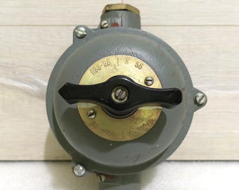 Op de afbeelding: Een grijs elektrisch component in industriële stijl met een zwarte handgreep en messing fittingen. Het ronde component heeft zichtbare schroeven en tekst, waaronder "ПВ3-25" en "56".