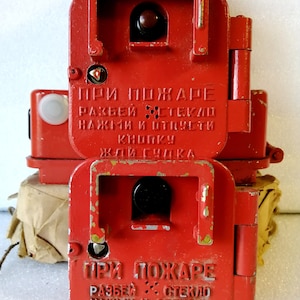 May include: Two vintage red fire alarm boxes stacked on top of each other. Each box has a rectangular shape with a hinged front panel. The text "ПРИ ПОЖАРЕ" is visible on the front, along with other Cyrillic text.