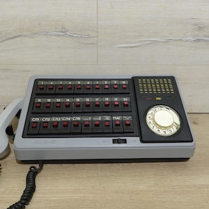 Op de afbeelding: Een grijze vintage draaitelefoon met een zwarte wijzerplaat en een toetsenbord met rode knoppen. De telefoon heeft een witte hoorn en een opgerold snoer.