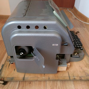 May include: A vintage gray metal typewriter with a paper holder and a keyboard. The typewriter is sitting on a wooden surface.