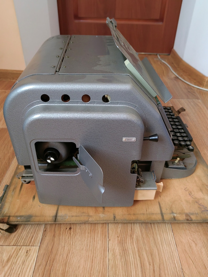 May include: A vintage gray metal typewriter with a paper holder and a keyboard. The typewriter is sitting on a wooden surface.
