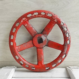 以下が含まれることがあります： 中央に正方形の穴が開いた、5本のスポークを持つ赤い金属製の車輪。車輪は使い古したような風合いがあり、小さな穴の輪で囲まれています。