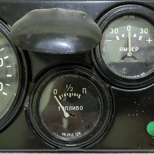 May include: A black dashboard with three round gauges. The gauges are labeled "AMEP", "Топливо", and "1/2". The gauge labeled "AMEP" has a needle pointing to 0. The gauge labeled "Топливо" has a needle pointing to 1/2. The gauge labeled "1/2" has a needle pointing to 0. There is a green light on the dashboard.