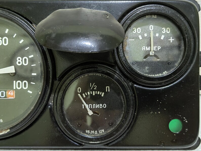 May include: A black dashboard with three round gauges. The gauges are labeled "AMEP", "Топливо", and "1/2". The gauge labeled "AMEP" has a needle pointing to 0. The gauge labeled "Топливо" has a needle pointing to 1/2. The gauge labeled "1/2" has a needle pointing to 0. There is a green light on the dashboard.