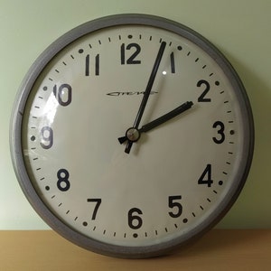 Puede incluir: Un reloj redondo gris con agujas negras y números negros. La esfera del reloj es blanca y tiene un borde negro. El reloj está puesto a las 11:55.