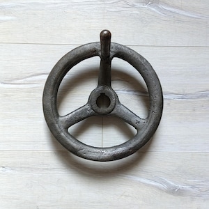 Könnte beinhalten: Ein großes, schwarzes Metallrad mit drei Speichen und einem Griff. Das Rad hat ein Loch in der Mitte und ist zum Drehen oder Bedienen einer Maschine gedacht.