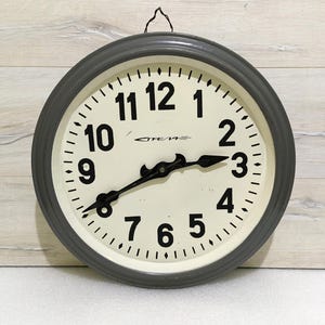 Può includere: Un orologio da parete vintage grigio con numeri e lancette neri. Il quadrante dell'orologio è bianco con un bordo nero e la parola "OTEMA" stampata in nero.