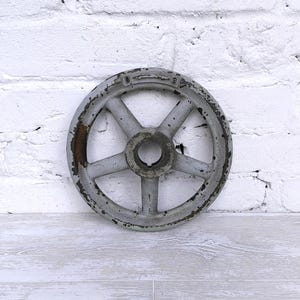Puede incluir: Un volante metálico gris de forma circular con cinco radios y un cubo central con chavetero. La superficie muestra signos de desgaste con pintura desconchada y manchas de óxido. El borde exterior presenta marcas en relieve.