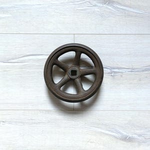 Könnte beinhalten: Ein rostiges Gusseisenrad mit sechs Speichen und einer quadratischen Nabe. Das Rad liegt auf einem hellgrauen Hintergrund mit Holzmaserung. Das verwitterte Aussehen des Rades deutet darauf hin, dass es sich um ein antikes oder industrielles Vintage-Teil handelt.