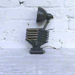 Könnte beinhalten: Industriell gestaltete, dunkelgraue, verstellbare Wandleuchte. Die Lampe hat einen Ziehharmonika-Arm und einen konischen Schirm. Die Lampe ist an einer weißen Backsteinmauer befestigt und hat ein sichtbares Netzkabel.