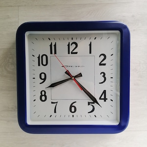 Könnte beinhalten: Eine quadratische Uhr mit blauem Rahmen, weißem Zifferblatt und schwarzen Zahlen. Die Zeiger der Uhr sind schwarz und rot, und die Uhr zeigt 4:05 Uhr an. Die Uhr hat das Wort "Time" in Schwarz auf dem Zifferblatt.