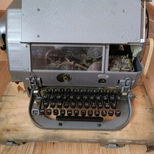 May include: A vintage gray typewriter with a black keyboard and Cyrillic lettering. The typewriter is on a wooden surface.