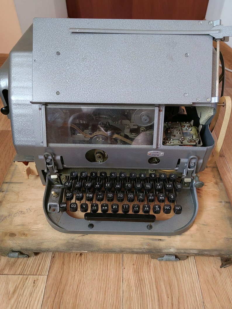 May include: A vintage gray typewriter with a black keyboard and Cyrillic lettering. The typewriter is on a wooden surface.