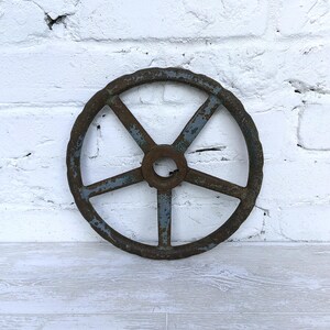 May include: A circular metal handwheel featuring five spokes radiating from a central hub. The object displays a weathered appearance with patches of blue paint peeling away to reveal a dark, rusted brown surface underneath the finish.