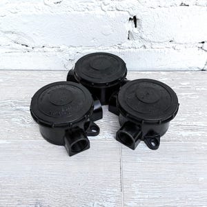 Puede incluir: Tres cajas de conexiones eléctricas negras con tapas redondas y aberturas laterales. Las cajas están hechas de un material duradero y están diseñadas para el cableado eléctrico. Están dispuestas sobre una superficie de madera clara contra una pared de ladrillos blancos.