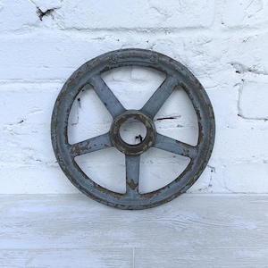 May include: A circular, five-spoke metal wheel with a weathered, grey-blue finish and visible patches of brown rust. The industrial component features a central hub with a hollow opening and a textured, worn surface throughout its entire structure.