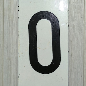May include: White metal sign with a black number zero. The sign has two holes at the top and bottom.