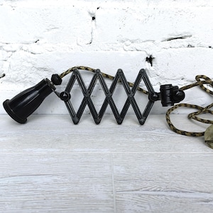 Könnte beinhalten: Schwarze Ziehharmonika-Lampenfassung mit schwarzer Glühbirnenfassung und einem Tarnmusterkabel. Die Lampe hat ein rautenförmiges Design und eine Vintage-Ästhetik. Der Stecker ist hellgrün.