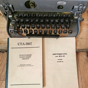 May include: A vintage teletype machine with a gray metal body and a black keyboard. The machine has a clear plastic cover over the top, revealing the internal workings. The keyboard has keys labeled with Cyrillic letters. The machine is sitting on a wooden surface. The text on the paper reads "СТА-М67" and "ЭЛЕКТРОДВИГАТЕЛЬ типа ДТА-40 ПАСПОРТ 193.10.00 ПС".