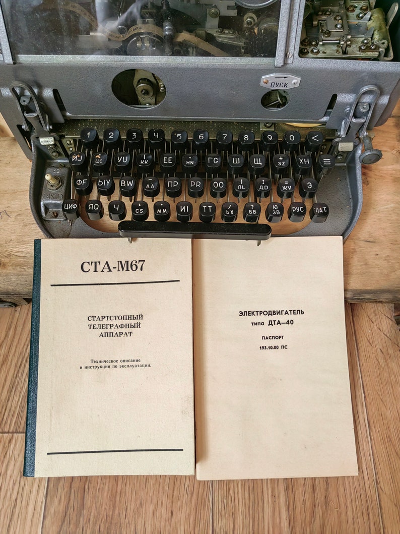 May include: A vintage teletype machine with a gray metal body and a black keyboard. The machine has a clear plastic cover over the top, revealing the internal workings. The keyboard has keys labeled with Cyrillic letters. The machine is sitting on a wooden surface. The text on the paper reads "СТА-М67" and "ЭЛЕКТРОДВИГАТЕЛЬ типа ДТА-40 ПАСПОРТ 193.10.00 ПС".