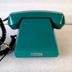 Puede incluir: Un teléfono rotatorio de color turquesa con un cable negro en espiral. El teléfono tiene una base cuadrada y un dial redondo.