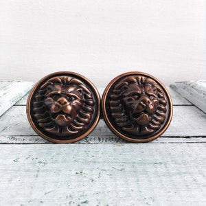 May include: Two round, copper-colored lion head drawer pulls with a detailed design.