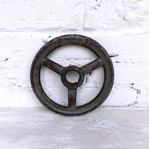 Pode incluir: Um volante manual de metal de três raios, de forma circular, com um acabamento cinzento-escuro desgastado e manchas de ferrugem visíveis. O cubo central apresenta uma abertura circular e o rebordo exterior mostra sinais de desgaste.