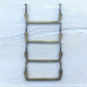 Peut inclure: Quatre poignées de tiroir en métal de couleur laiton disposées en pile verticale. Chaque poignée présente un design en barre rectangulaire avec deux boulons de montage filetés et des écrous fixés aux extrémités de la structure.
