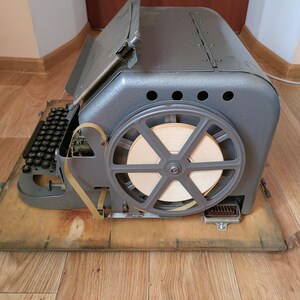 May include: A vintage gray metal typewriter with a paper tape spool and a keyboard. The typewriter is sitting on a wooden surface.