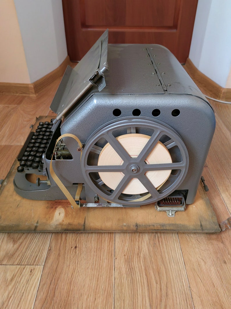 May include: A vintage gray metal typewriter with a paper tape spool and a keyboard. The typewriter is sitting on a wooden surface.