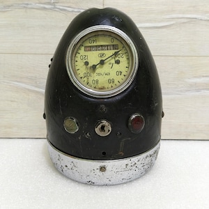 Peut inclure: Un compteur de vitesse de moto vintage noir avec une lunette chromée. Le compteur de vitesse a un cadran blanc avec des chiffres noirs et une aiguille jaune. Le compteur de vitesse est logé dans un boîtier en forme d'œuf avec un revêtement en cuir noir. Le boîtier a un trou de serrure et plusieurs petits voyants.