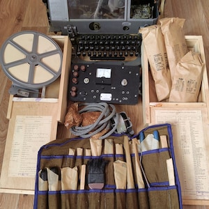 May include: A vintage, gray metal typewriter with a keyboard, a reel of film, and a tool roll with various tools. The typewriter is open and the keyboard is visible. The tool roll is brown with blue trim and contains a variety of tools, including screwdrivers, pliers, and a brush. The tools are wrapped in brown paper.