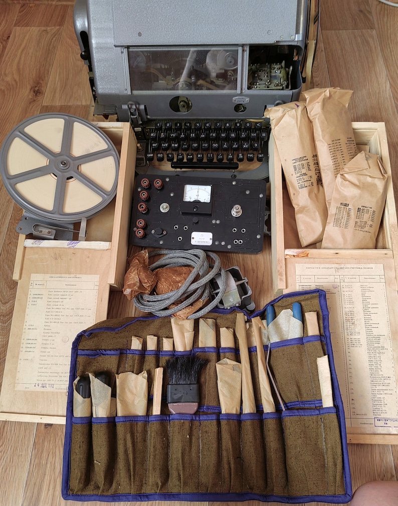 May include: A vintage, gray metal typewriter with a keyboard, a reel of film, and a tool roll with various tools. The typewriter is open and the keyboard is visible. The tool roll is brown with blue trim and contains a variety of tools, including screwdrivers, pliers, and a brush. The tools are wrapped in brown paper.