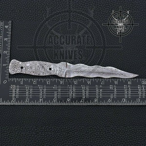 May include: A Damascus steel dagger with a wavy blade and handle. The knife is approximately 9 inches long, with a detailed pattern on the metal. The Accurate Knives logo is visible in the background.