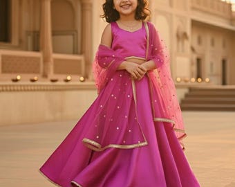 May include: A young girl in a magenta traditional Indian outfit, including a crop top, long skirt, and sheer scarf. The skirt has a layered design with gold trim. The outfit is worn with a small headpiece. The background is a building with arches.