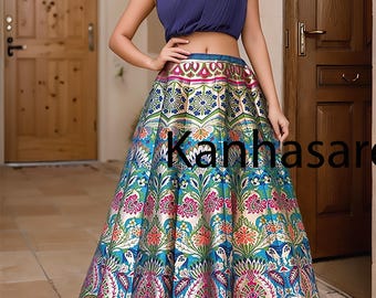 May include: A woman wearing a blue crop top and a long, colorful skirt with a floral pattern. The skirt has a silver metallic sheen and a pink and green border. The woman is standing in front of a wooden door.