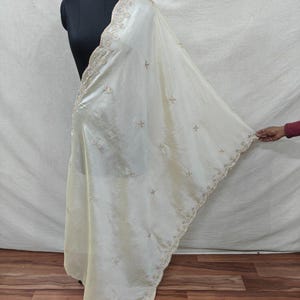 May include: Cream-coloured silk shawl with detailed gold embroidery along the borders and across the surface. The shawl is draped over a black mannequin, highlighting its flowing design and delicate features. The embroidery includes floral and geometric patterns.