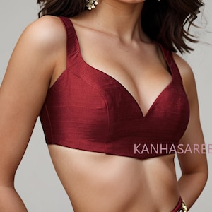 Peut inclure: Un haut de culture court, sans manches, de couleur bordeaux, avec un décolleté en V profond et un corsage ajusté. Le haut est fait d'un tissu lisse et soyeux. Le texte "KANHASARE" est visible dans le coin inférieur droit de l'image.