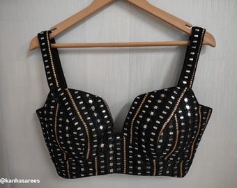 May include: Black sequined top with square and vertical patterns, hanging on a wooden hanger. The top has thick straps and a deep V-neckline. The design includes silver and gold accents.