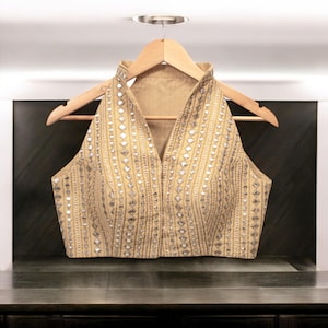 May include: A sleeveless, beige blouse with a high collar and a V-neckline. The blouse is decorated with silver sequins and mirror work in a geometric pattern.