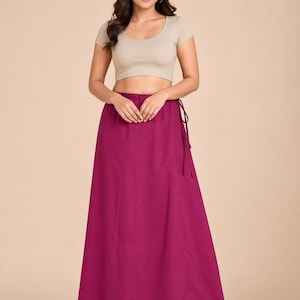 May include: A woman wearing a beige crop top and a long, flowing magenta skirt. The skirt is tied at the waist with a matching drawstring. The outfit is simple and elegant, suitable for various occasions.