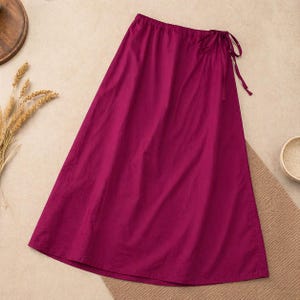May include: A long, magenta skirt with a drawstring waist and tie closure. The skirt is made of a lightweight fabric and has a flowing silhouette. The skirt is a solid color and is suitable for casual wear.